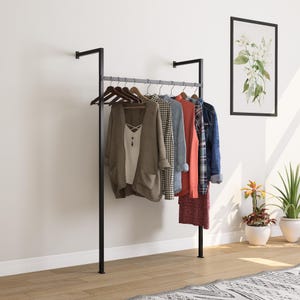 Puede incluir: Un perchero de metal negro con una barra horizontal y soportes verticales, que sostiene varias prendas en perchas de madera. La ropa incluye una rebeca marrón, una camiseta blanca, una camisa a cuadros, una camisa roja y una chaqueta vaquera. Una impresión floral enmarcada cuelga de la pared.