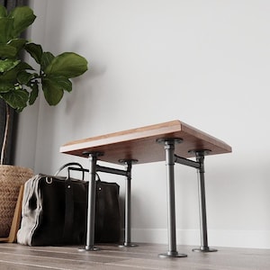 Puede incluir: Banco rectangular de madera oscura, sostenido por patas de tubo metálico negro. El banco está contra una pared blanca, con una bolsa gris oscuro grande y una planta en maceta en el fondo. El banco mide unos 91 cm de largo.