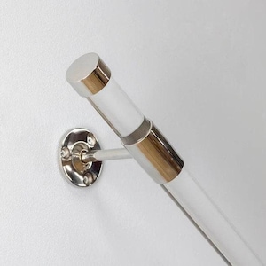 Könnte beinhalten: Ein moderner Handlauf mit einem transparenten Acrylrohr und polierten Chromakzenten. Der Handlauf ist an einer weißen Wand mit einer kreisförmigen Chrom-Wandhalterung befestigt. Die Endkappe ist ebenfalls verchromt.
