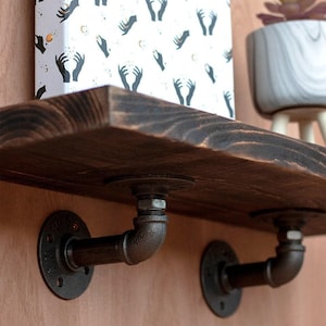 Peut inclure: Étagère en bois foncé avec des supports de tuyaux industriels. L'étagère supporte une boîte décorative avec un motif de mains tenant des étoiles et une petite plante en pot. Les supports sont en métal noir.