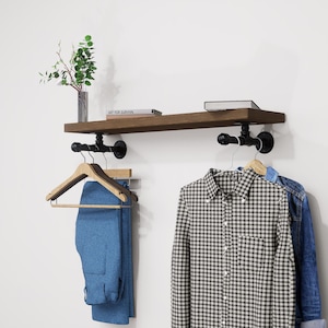 Op de afbeelding: Een rustiek houten plank met zwarte pijpfittingen, met een boek en een kleine vaas met groen. Daaronder hangen een blauwe broek en een geruit overhemd aan houten kleerhangers. Een spijkerjasje is ook zichtbaar.