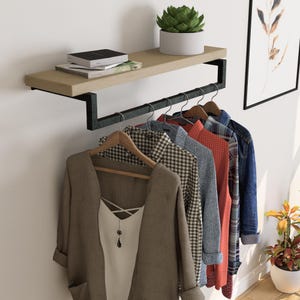 Puede incluir: Un perchero de pared con una estantería de madera clara y una barra de metal negro para colgar ropa. Varias camisas y una chaqueta cuelgan de perchas de madera. Una planta suculenta en una maceta blanca se encuentra en la estantería, con libros y una impresión decorativa.