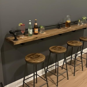 Puede incluir: Un bar rústico con una estantería de madera con soportes de tubería negra y taburetes a juego. La estantería contiene varias bebidas y vasos. Los taburetes tienen patas de metal y asientos de madera con un logotipo.