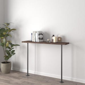 May include: A dark brown wooden console table with two dark grey metal legs. The table is against a white wall and holds books, candles, and perfume bottles. A potted plant is to the left.
