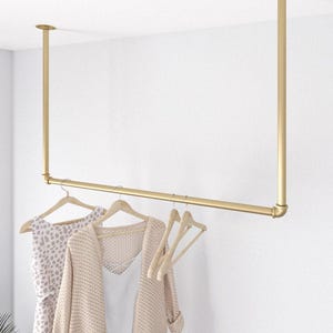 May include: A gold-colored, ceiling-mounted clothing rack. The rack is made of metal pipes and has a horizontal bar for hanging clothes. Several wooden hangers hold a dress and a sweater. The rack is mounted against a white wall.