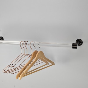 May include: A clear acrylic clothing rod mounted on a white wall with black hardware. Several hangers are displayed, including copper-colored metal and natural wood hangers. The rod is designed for organizing clothing.