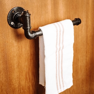 Può includere: Portasciugamani in tubi industriali neri montato su una superficie di legno. Un asciugamano bianco con tre strisce beige è appeso. Il portasciugamani è realizzato con tubi e raccordi in metallo nero.