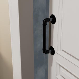 May include: Black industrial-style door pull handle made from pipes and fittings. The handle is mounted on a white door and a gray wall. The handle is a vertical bar with two circular mounting plates.