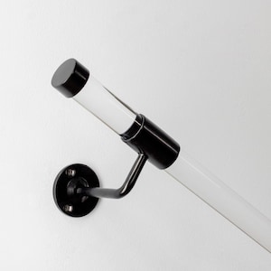 Pode incluir: Um corrimão moderno com uma barra cilíndrica transparente e detalhes em metal preto. O corrimão é montado em uma parede branca com uma base preta circular. Design minimalista e contemporâneo.