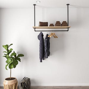 Puede incluir: Un perchero moderno de estilo industrial con una estantería de madera y una barra para colgar. El perchero está suspendido del techo con tuberías negras. Un suéter azul oscuro y una chaqueta negra cuelgan de perchas de madera. Una bolsa de cuero marrón y toallas están en el estante.