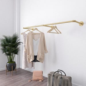 Pode incluir: Um varão de roupa de metal dourado montado numa parede branca, com um cardigan bege, um top branco e um vestido estampado. Uma mala rosa e uma mala de viagem cinzenta estão no chão de madeira. Uma planta em vaso está no canto.