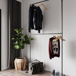 May include: A black metal clothing rack with two horizontal bars. A black leather jacket and a dark blue sweater hang on the top bar. A burgundy sweater with a graphic and a gray duffel bag sit below. A potted plant is to the left.
