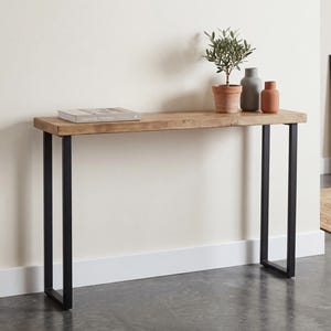 Puede incluir: Una consola de madera con acabado natural y patas de metal negro. La mesa está decorada con una pequeña planta en maceta, dos jarrones y un libro. La mesa mide 122 cm de largo.