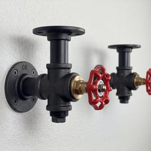 May include: Two industrial-style wall-mounted pipe fittings. The fittings are black with red valve handles and brass accents. The fixtures are mounted on a white wall, showcasing a rustic, functional design.