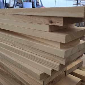May include: A pile of untreated wooden planks, displaying the natural grain and light brown hue of the timber. The rectangular planks look freshly sawn, suitable for building or woodworking tasks.