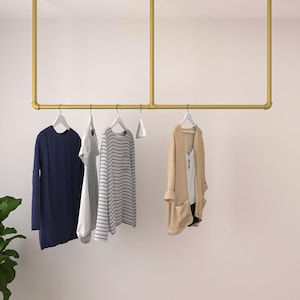 May include: A gold-colored, ceiling-mounted clothing rack with a horizontal bar and vertical supports. Several garments hang on white hangers, including a navy blue long-sleeve top, a white dress, a striped shirt, and a beige cardigan.