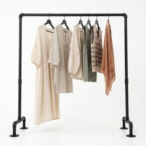 Peut inclure: Un portant à vêtements en tuyaux industriels noirs supporte six pièces sur des cintres noirs. On y trouve une longue robe crème, une tunique beige, un chemisier clair, un haut sans manches vert sauge, un gilet en tricot et une écharpe marron.