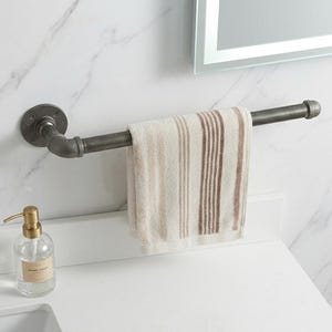 Puede incluir: Un toallero de estilo industrial hecho de tuberías de metal gris oscuro. Una toalla a rayas beige y marrón cuelga de la barra. Un dispensador de jabón de vidrio transparente se encuentra en la encimera blanca.