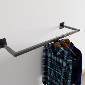 Puede incluir: Un perchero de metal gris oscuro montado en una pared blanca. El perchero sostiene tres perchas de madera con una camisa a cuadros azul y una chaqueta vaquera. El perchero tiene un diseño simple y moderno.