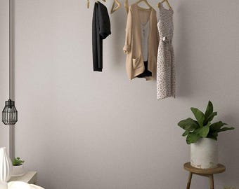 May include: A black metal clothing rack suspended from the ceiling, holding a black shirt, a tan cardigan, and a patterned dress. The room has a neutral color palette with a bed and a small side table with a plant.