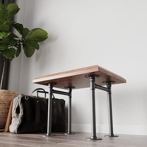 Puede incluir: Banco rectangular de madera marrón oscuro, sostenido por tuberías metálicas negras. El banco mide aproximadamente 122 cm de largo y 41 cm de ancho. El diseño industrial presenta conexiones de tuberías visibles y una construcción robusta.