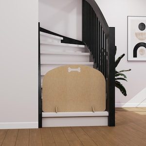 Op de afbeelding: Een lichtbruine huisdierpoort met een botvormige uitsparing aan de bovenkant, ontworpen om trappen te blokkeren. De poort is gemaakt van lichtgekleurd hout en heeft twee kleine voetjes voor stabiliteit. De trap heeft witte treden en een zwarte leuning.