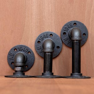 Puede incluir: Tres accesorios de tubería industriales negros, montados sobre madera. Cada uno presenta una placa circular con el texto "1949" y una sección de tubería curvada. Los accesorios son de diferentes alturas, posiblemente para estanterías o decoración.