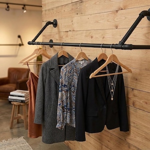 Puede incluir: Un perchero de tubería metálica negra montado en una pared de madera. Varias prendas cuelgan de perchas de madera, incluyendo un abrigo gris, una blusa estampada y una blazer negra. El perchero está en un entorno de boutique.