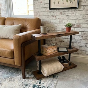 May include: A three-tiered, rustic side table with dark wood shelves and black pipe supports. The table holds a mug, books, a tablet, a blanket, and a basket with remote controls. The table is next to a brown leather sofa.