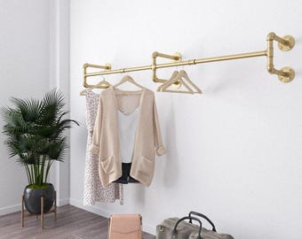 May include: A gold-colored metal clothing rack mounted on a white wall, holding a variety of garments on wooden hangers. A potted plant, a purse, and a large gray duffel bag are on the floor.