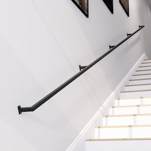 Puede incluir: Una barandilla de metal negro instalada en una pared blanca, en ángulo para seguir la escalera. La barandilla está soportada por varios soportes negros. Las escaleras son blancas con peldaños de madera clara. Obras de arte cuelgan encima de la barandilla.