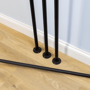 Puede incluir: Patas de muebles de tubo de metal negro con bridas redondas. Las patas están sobre un suelo de madera clara junto a un rodapié blanco y una pared azul claro. La imagen muestra las patas y un tubo horizontal.