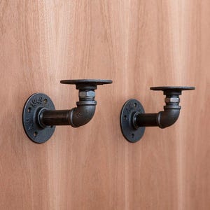 Puede incluir: Dos soportes de estantería industriales negros de tubería. Cada soporte tiene una base redonda con el texto "1949", una tubería curvada y una parte superior plana y circular. Los soportes están montados sobre una superficie de madera.