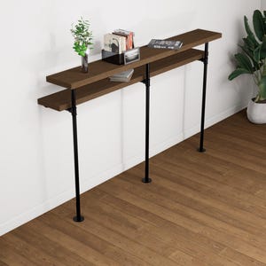 Peut inclure: Une console en bois brun foncé à deux niveaux avec des pieds en tuyaux métalliques noirs. La table est contre un mur blanc et comporte des objets décoratifs sur l'étagère supérieure, dont des livres et une petite plante.