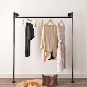Pode incluir: Um cabideiro de metal preto exibe um par de calças cinza escuro, um cardigan bege e um vestido estampado. Uma bolsa de couro marrom, sandálias e um tapete redondo estão no chão. O cabideiro está contra uma parede branca e um chão de madeira.