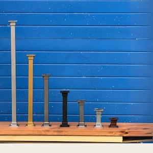Peut inclure: Une collection de colonnes décoratives de différentes hauteurs et couleurs, dont argent, or, gris, noir et marron. Les colonnes sont exposées sur une surface en bois sur fond bleu.