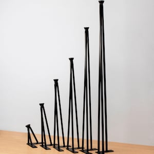 May include: A collection of seven black metal hairpin table legs, each of a different height, arranged in a stepped sequence. Each leg features a flat base and a rounded top. The legs are displayed on a light wood surface against a white backdrop.