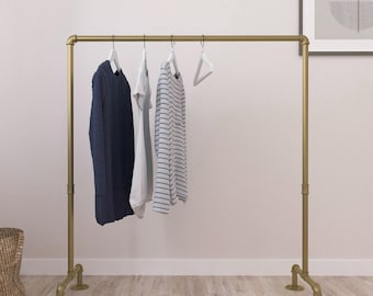 May include: A gold-colored clothing rack with a minimalist design. The rack holds a navy blue sweater, a white t-shirt, and a striped shirt on white hangers. The rack is made of metal pipes and has a sturdy base.