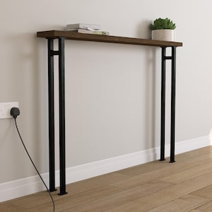 Może przedstawiać: Konsola z ciemnego drewna z czarnymi metalowymi nogami. Stół ma prostokątny blat i minimalistyczny design. Mała roślina doniczkowa i książki znajdują się na stole. Stół stoi przy białej ścianie.