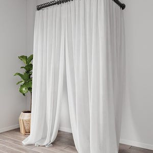 Könnte beinhalten: Weiße, transparente Vorhänge hängen an einer schwarzen Stange und bilden einen Raumteiler. Die bodenlangen Vorhänge sind leicht gerafft, sodass Licht durchscheinen kann. Eine Topfpflanze in einem geflochtenen Korb steht in der Ecke und sorgt für einen Hauch von Grün.