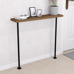 Peut inclure: Une console en bois marron avec des pieds en métal noir. La table est ornée de livres, de deux photos encadrées et d'une plante en pot avec des fleurs roses. La table est placée contre un mur blanc.