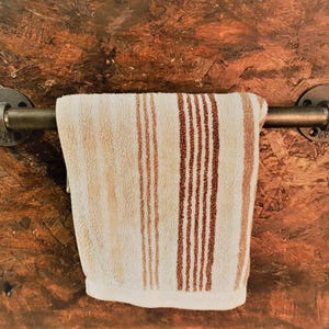 Puede incluir: Un toallero rústico hecho de tuberías metálicas, montado sobre una superficie marrón texturizada. Una toalla beige con rayas verticales marrones cuelga del toallero, creando una estética cálida e industrial. Adecuado para baño o cocina.