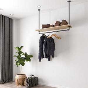 Può includere: Un attaccapanni moderno in stile industriale sospeso al soffitto. Presenta una mensola in legno con asciugamani marroni piegati e una pochette in pelle. Sotto, una giacca nera e un maglione blu scuro sono appesi a grucce di legno. Una grande pianta e una borsa da viaggio sono sul pavimento.