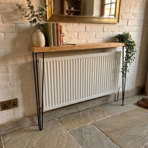 Pode incluir: Uma consola rústica com tampo de madeira natural e pernas de hairpin pretas. A mesa está posicionada em frente a um radiador branco. Inclui livros, um vaso e vegetação. Um espelho está pendurado acima da mesa.