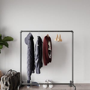 Puede incluir: Un perchero de metal negro con varias prendas colgadas. Incluye una bufanda de punto gris oscuro, una chaqueta de cuero negra y un suéter burdeos. Zapatillas blancas y una bolsa de lona gris en la base.