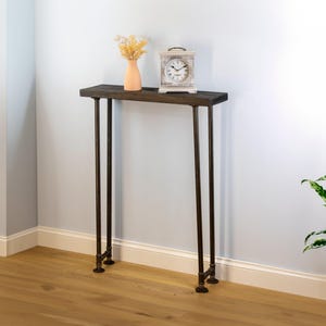 May include: A dark wood console table with metal legs. A peach-colored vase with dried flowers and a vintage clock sit on the table. The table is against a light blue wall with a wooden floor.