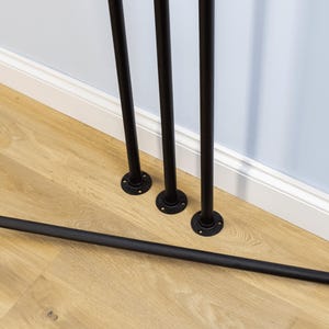 May include: Black metal pipes with circular flanges, arranged on a light wood floor. Three pipes stand upright, while a fourth lies horizontally. The pipes are near a white baseboard and a light blue wall.
