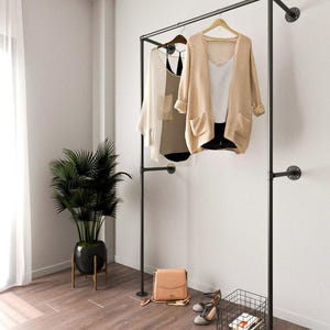 May include: A black metal clothes rail with two tops hanging on wooden hangers, a beige cardigan and a white top. A tan handbag, grey heels, and a black wire basket with books are on the floor. A potted plant is to the left.
