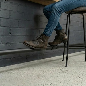 May include: A black metal bar stool with a wooden seat and a black pipe footrest. The image shows a person wearing jeans and brown boots, resting their foot on the pipe. The stool is set against a grey brick wall.