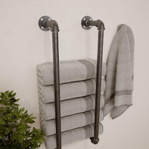 Peut inclure: Porte-serviettes de style industriel en tuyaux métalliques foncés, monté sur un mur clair. Il contient une pile de serviettes grises et blanches rayées, avec une serviette suspendue sur le côté. Design fonctionnel et décoratif.